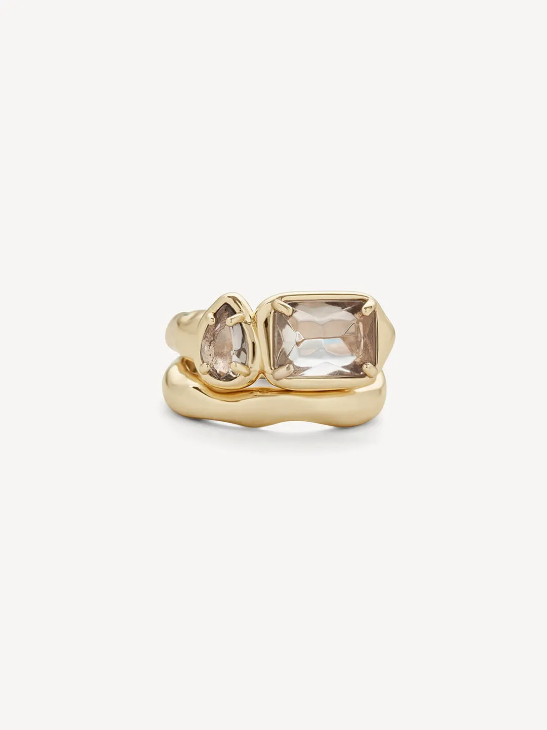 A gold ring featuring a large rectangular clear gemstone and a smaller teardrop-shaped clear gemstone, set in a polished gold band against a light gray background.